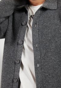 Graue Strickjacke mit strukturierter Oberfläche, ausgestattet mit einem Kragen, sechs Knöpfen und einer lockeren Passform über einem weißen T-Shirt darunter.