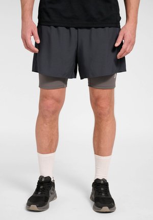 Man draagt zwarte sportshorts over grijze compressieshorts, witte sportsokken en zwarte hardloopschoenen, staand op een witte achtergrond.