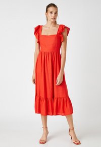 Vestido de lino rojo con escote cuadrado, mangas acampanadas y dobladillo en capas. Modelo combinado con tacones rojos con tiras. Textura suave.