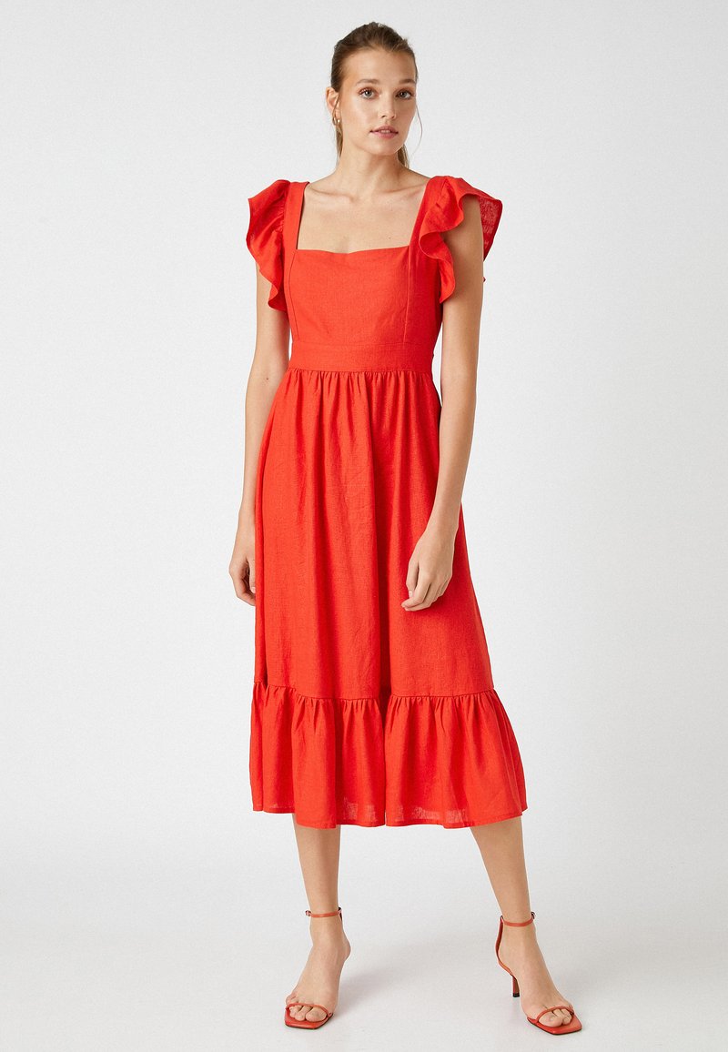 Vestido de lino rojo con escote cuadrado, mangas acampanadas y dobladillo en capas. Modelo combinado con tacones rojos con tiras. Textura suave.