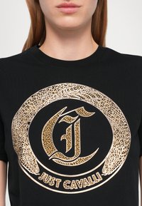 Schwarzes T-Shirt mit einem großen goldenen Grafikmotiv, das komplizierte Muster zeigt und den Text "Just Cavalli" am unteren Rand trägt, aus weichem Baumwollmaterial gefertigt.