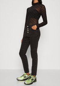 Top negro semitransparente de manga larga con textura estampada. Jeans negros de cintura alta con cinco botones y acentos acanalados en las piernas. Zapatillas plateadas y verde neón.