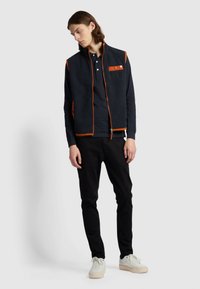 Marine fleece vest met oranje accenten en rits, gedragen over een donker shirt. Slim-fit zwarte broek en witte sneakers maken de outfit compleet.