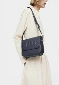 Bolso de hombro de cuero azul marino con el logo "TOUS" en relieve, forma rectangular, cierre con solapa, correa ajustable, textura suave.