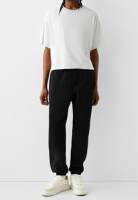 Vit, kortärmad, croppad t-shirt i kombination med svarta, löst sittande sweatpants och krämfärgade sneakers, stående på en ljus bakgrund.