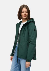 Giacca verde con cappuccio e chiusura a zip, realizzata in materiale liscio. Presenta tasche frontali e un'etichetta nera con logo sulla manica.