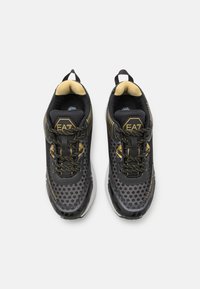 Schwarze Sportschuhe mit einem strukturierten Mesh-Obermaterial, goldenen Akzenten und dunklen Schnürsenkeln. Verfügt über eine gepolsterte Sohle und eine Zuglasche an der Ferse.