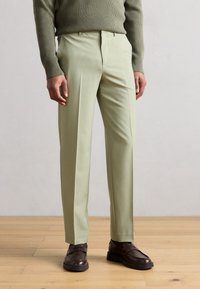 Lysegrønne skræddersyede bukser med en glat tekstur og kantdetalje, kombineret med mørkebrune slip-on sko og en ribbet olivengrøn sweater.