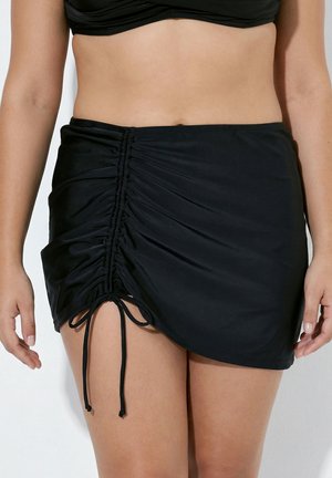 Vrouw draagt een zwart geplooid mini rokje met verstelbaar trekkoord en een bijpassend zwart cropped topje, zichtbaar van de taille tot halverwege het bovenbeen.