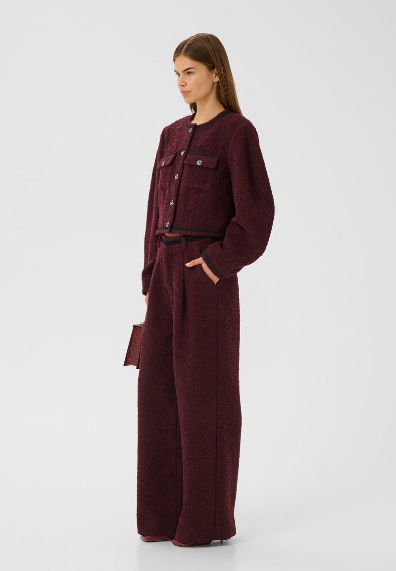 Burgunderfarbene Texturjacke mit zwei Fronttaschen, silbernen Knöpfen und einer passenden hochtaillierten, weiten Hose. Inklusive einer gemusterten Handtasche.