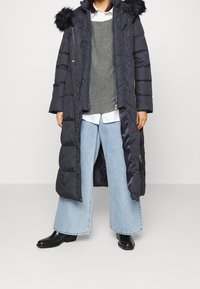 Lange marineblauwe donzen jas met bontgevoerde capuchon, ritssluiting, steekzakken aan de voorkant, gedragen over een grijze gebreide top en wijde blauwe jeans. Zwarte enkellaarzen.