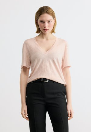 Frau mit kurzen blonden Haaren, die goldene Creolen, ein hellrosa V-Ausschnitt-T-Shirt, schwarzen Gürtel und schwarze Hose trägt, stehend vor weißem Hintergrund.