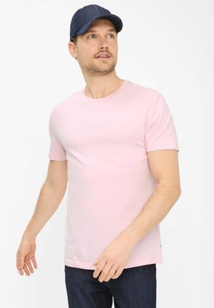 Man draagt een lichtroze shirt met korte mouwen, donkere spijkerbroek en een donkerblauwe baseballpet, kijkt omhoog naar rechts.