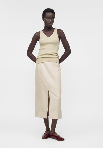 Γυναίκα στέκεται με τα χέρια πίσω από την πλάτη, φοράει εκρού ριμππλέ αμάνικο τοπ, μπεζ μίντι φούστα με εμπρόσθια σχισμή και σκούρα κόκκινα παπούτσια Mary Jane.