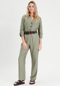 Olivgrüner Jumpsuit mit durchgehender Knopfleiste, hochrollbaren Ärmeln, zwei Taschen und einem schwarzen Gürtel mit einer runden Holzschnalle. Schwarze Sandalen.