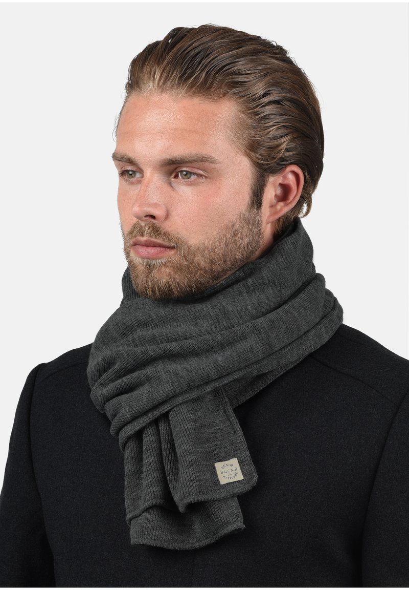 Tumši pelēks adīts šalle ar ribotu tekstūru un nelielu etiķeti netālu no gala. Valdā brīvi ap kaklu pār mēteļa.