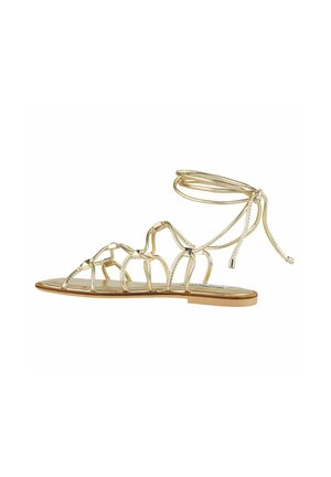 Sandali piatti metallici oro con cinturini intrecciati e una cavigliera legata. Sottopiede liscio con design minimalista e suola piatta.