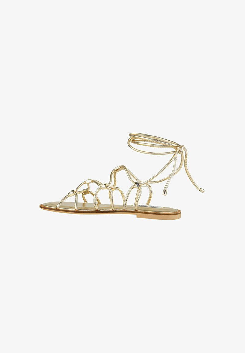 Sandali piatti metallici oro con cinturini intrecciati e una cavigliera legata. Sottopiede liscio con design minimalista e suola piatta.