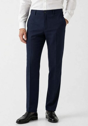 Uomo che indossa pantaloni blu navy su misura abbinati a mocassini in pelle neri e una camicia bianca, in piedi con le mani nelle tasche.