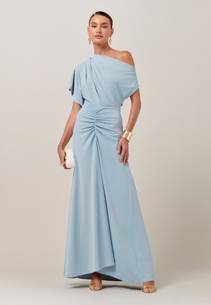 Naine helesinise üheõlalise voltidega kleidiga, hoides valget käekotti, kandes kuldseid kõrvarõngaid, käevõru ja avatud varbaga kontsakingi neutraalsel taustal.