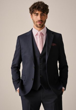 Costume marine avec revers crantés, gilet et pantalons assortis. Chemise à carreaux roses et cravate, avec une pochette de poitrine. Tissu texturé.