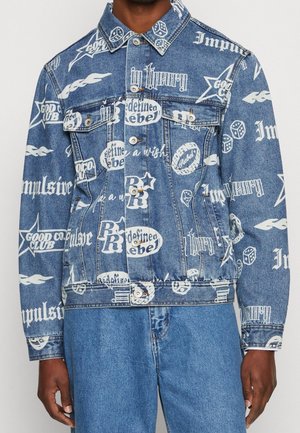Jeansjacke mit weißem grafischem Text und Symbolen einschließlich "rebel", Sternen, Würfeln und Flammen, getragen zu passenden Jeans.