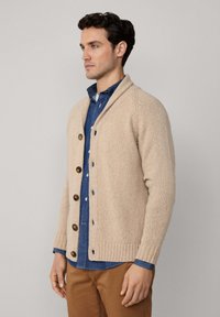 Man in een beige knopenvest over een blauwe shirt en een bruin broek, staande zijwaarts tegen een effen lichte achtergrond.