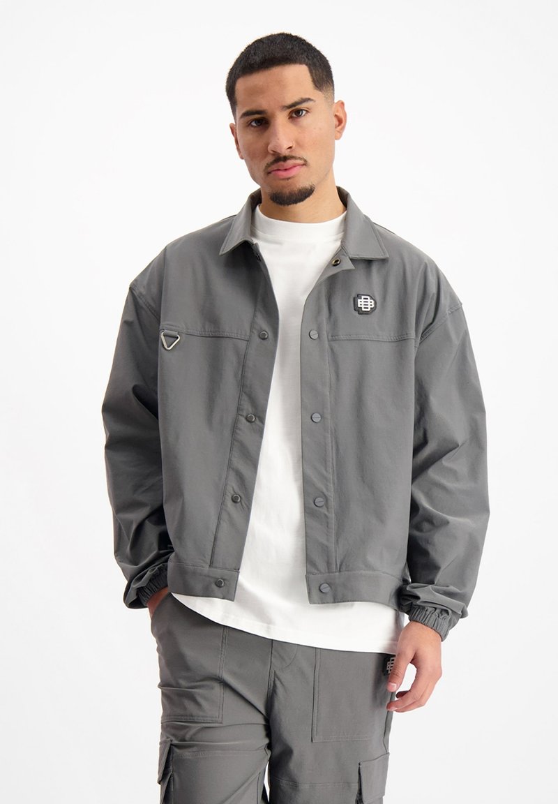 Grijze button-up jas met een ontspannen ontwerp, voorzien van een logo patch, opvallende kraag en elastische manchetten. Draagt over een wit shirt.