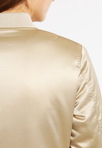 Giacca bomber in raso dorato con colletto a costine, texture liscia e cuciture sulle spalle che mostrano delle leggere pieghe per una maggiore dimensione.