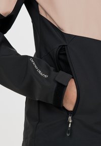 Svart och tan mjukskal jacka med en dragkedjeficka, justerbar manschett med Velcro och en slät textur på tyget.