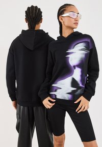 Svart hettegenser med et lilla og hvitt forvrengt grafisk design på forsiden, med ribbede mansjetter og nederkant, brukt sammen med svarte shorts.