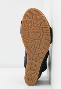 Semelle de chaussure noire avec un design en caoutchouc texturé. Présente un logo Timberland bien en vue et un motif de bande de roulement marron clair pour l'adhérence.
