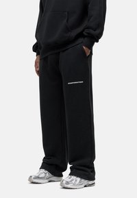 Schwarzer oversized Hoodie mit Fronttasche und passenden schwarzen Jogginghosen, die weißen Text "GOODFORNOTHING" aufweisen. Silbergraue Sportschuhe mit Netzstruktur.