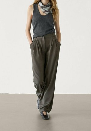 Woman wearing a dark gray sleeveless top, loose olive-green trousers, black shoes, and a plaid neck scarf walking with hands in pockets.