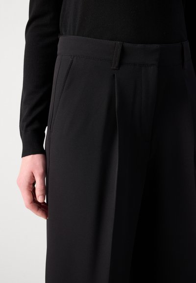 Pantalon noir sur mesure avec un devant plat, des poches latérales et une texture lisse. La taille se situe au-dessus des hanches.
