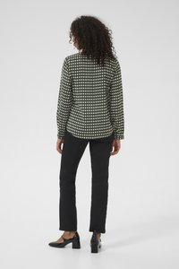 Woman with curly hair wearing a green patterned long-sleeve blouse, black pants, and black block-heeled shoes, standing facing away.