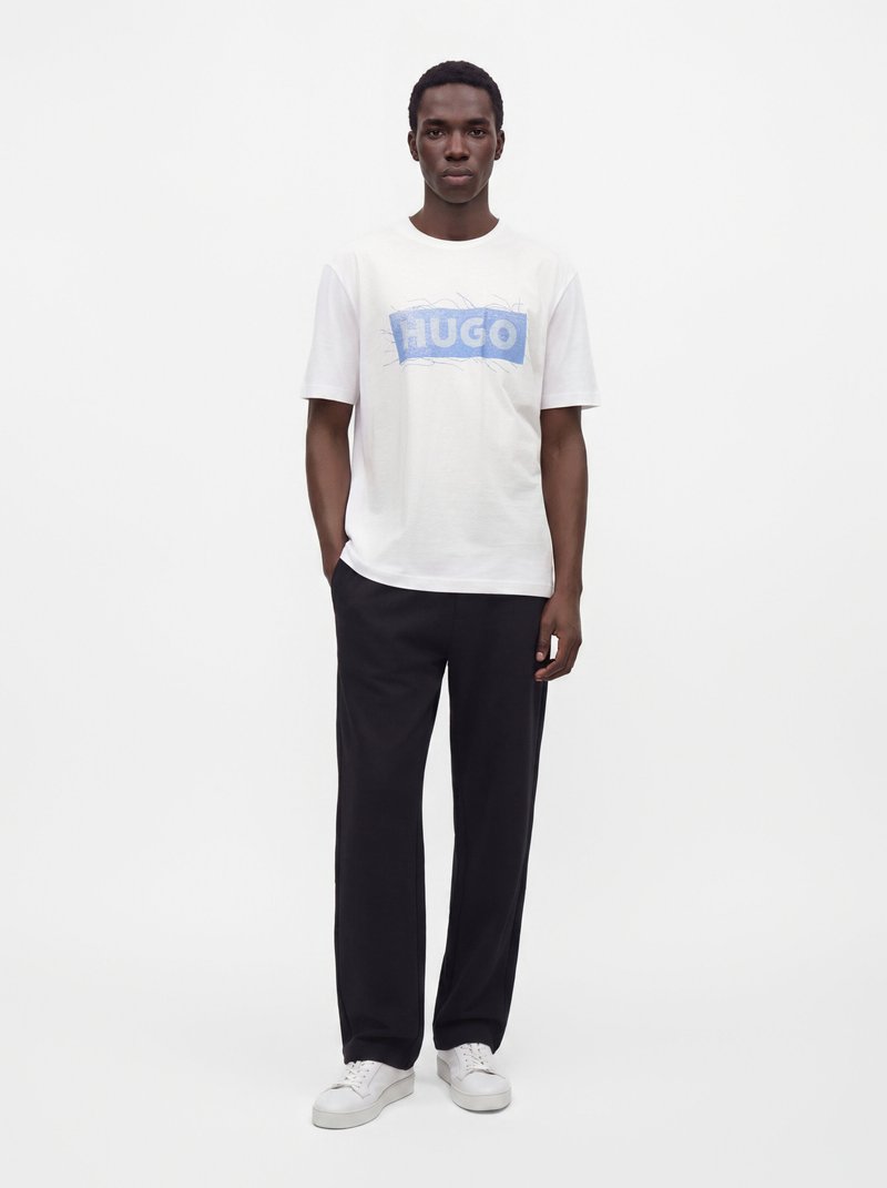 Млад мъж, облечен с бяла тениска с син текст "HUGO", черен панталон и бели маратонки на бял фон.