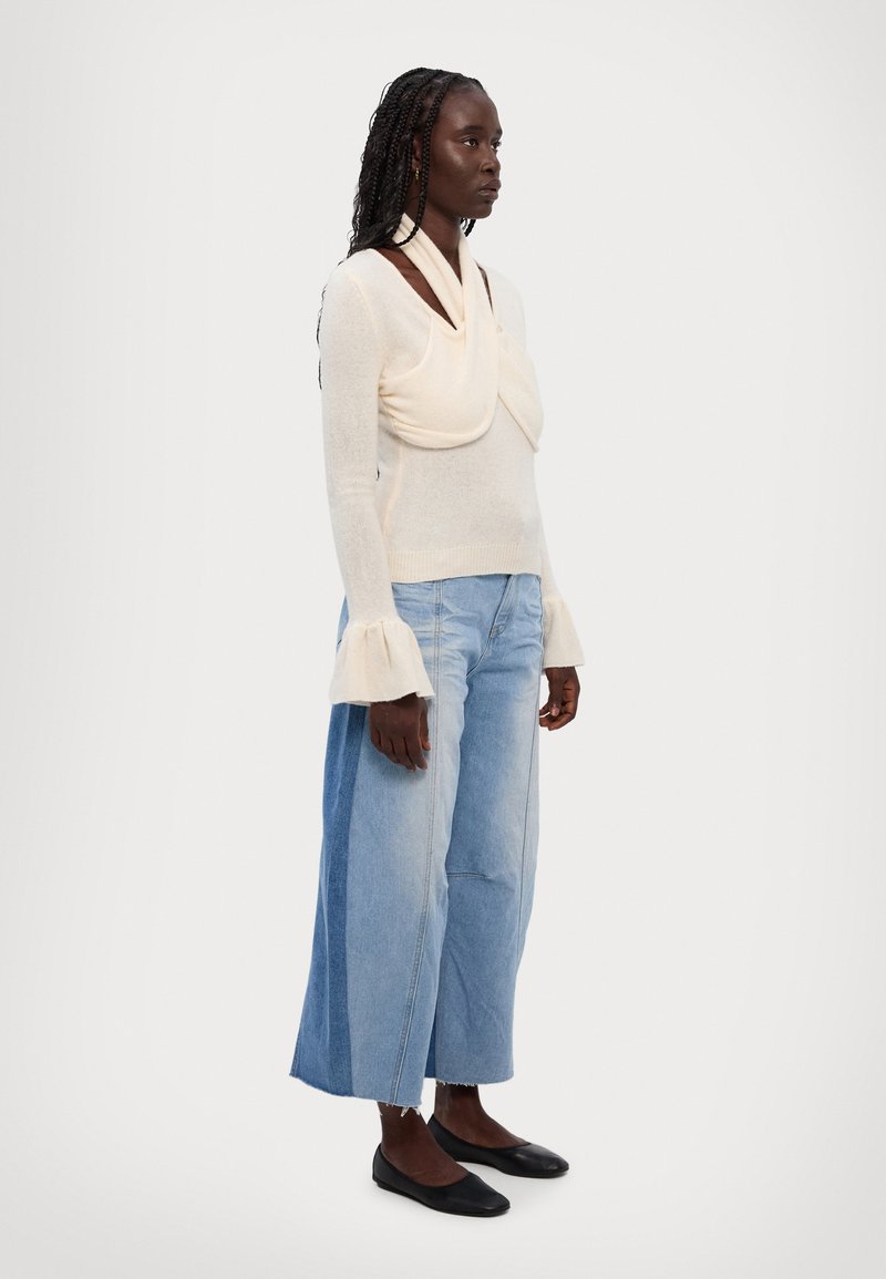 Maglietta a maniche lunghe in maglia beige chiaro con scollo drappeggiato e maniche a campana; abbinata a culotte di denim con spacco in diverse sfumature di blu e ballerine nere.