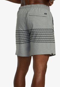 Mann trägt graue Shorts mit schwarzen, geometrischen horizontalen Streifen und Gesäßtasche, steht mit den Händen in der Hüfte, Beine sichtbar vom mittleren Oberschenkel abwärts.