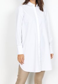 Camicia bianca con colletto, maniche lunghe e orlo curvo. Realizzata in un tessuto liscio, presenta un design fresco e pulito.