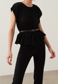 Haut noir plissé avec des manches courtes côtelées, corsage ajusté et basque évasée. Porté avec un pantalon noir slim et une fine ceinture noire. Texture lisse.