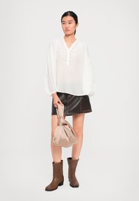Femme portant une blouse blanche, une minijupe en cuir brun foncé, des bottes marron mi-mollet, tenant un sac à main beige noué, se tenant contre un fond blanc.