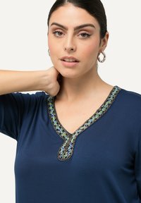 Námořnické modré tričko s véčkovým výstřihem, které má podrobenou výzdobu z korálků v zelené, modré a černé barvě podél výstřihu. Měkká textilie.