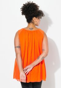 Top senza maniche di un brillante arancione con schiena arricciata, caratterizzato da una silhouette fluida e un tessuto leggero. Leggings neri con un motivo sottile.