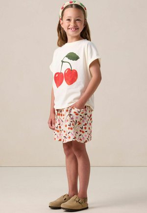 T-shirt blanc en coton avec un graphisme de cerise rouge, associé à un short crème avec un imprimé cerise coloré. Chaussures slip-on marron et un bandeau.