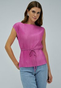 Mujer vestida con una blusa de punto rosa sin mangas con un cinturón atado, combinada con jeans azul claro, frente a un fondo gris liso.