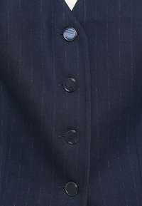 Granatowa marynarka w prążki z czterema okrągłymi guzikami, górny guzik z wygrawerowanym napisem "GUESS", a pod spodem widoczny biały kołnierzyk koszuli.