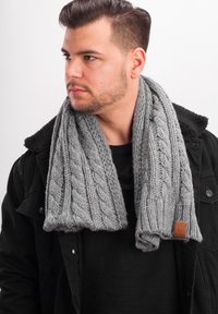 Gray knitted scarf featuring a prominent cable pattern and a textured finish, with a brown leather accent tag. Worn over a black sweater.