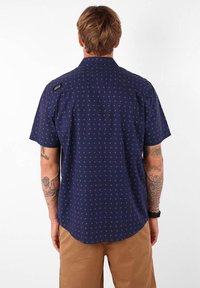Chemise à manches courtes, bleu marine, en tissu avec un motif géométrique dans des teintes plus claires. Dotée d'un col classique et d'une étiquette de marque sur l'épaule.