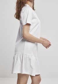 Žena v krátkých rukávech bílých šatech s nabíranou sukní stojící bokem na jednotném světlém pozadí.